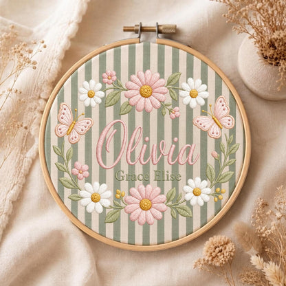 May include: A circular wooden embroidery hoop containing fabric with vertical sage green and white stripes. The embroidery features the name Olivia in pink script, the name Grace Elise in green text, pink daisies, white daisies, and two pink butterflies.