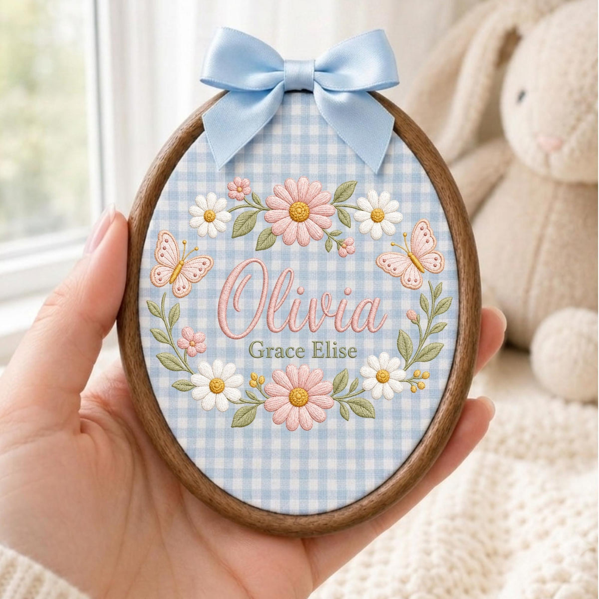 May include: An oval wooden embroidery hoop frame containing fabric with a light blue and white gingham pattern. The fabric features embroidered pink and white daisies, green leaves, two pink butterflies, and the names Olivia and Grace Elise.