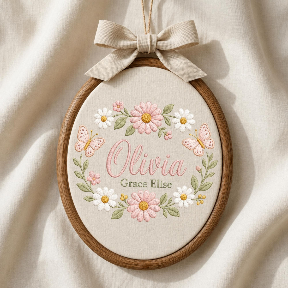 May include: An oval wooden embroidery hoop containing cream fabric with the embroidered name Olivia Grace Elise in pink and green. The text is surrounded by a floral wreath of pink and white daisies, green leaves, and two pink butterflies. A beige ribbon bow is attached to the top.