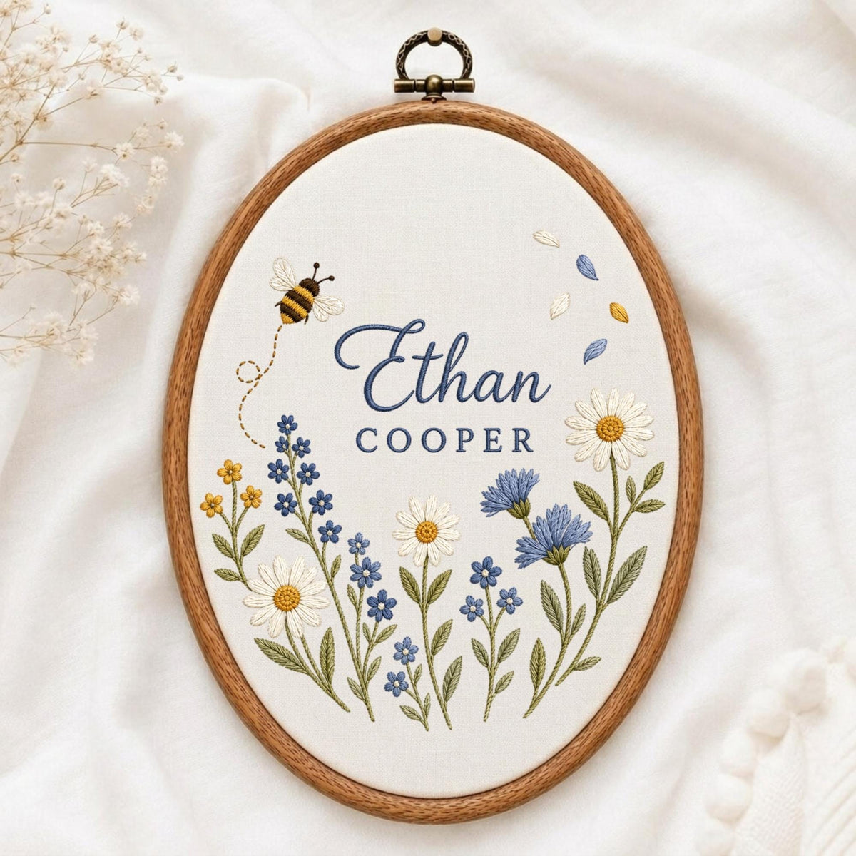 May include: An oval wooden embroidery hoop containing white fabric with the embroidered name Ethan Cooper in blue script and block letters. The design features a small bee, scattered petals, and a variety of blue and white wildflowers with green stems.