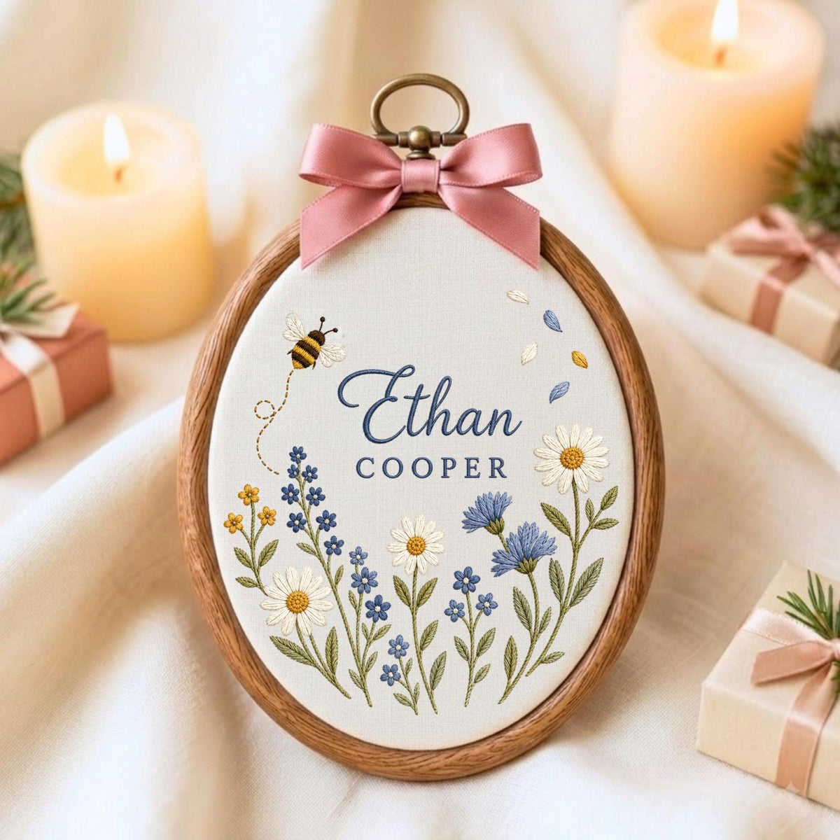 May include: An oval wooden embroidery hoop containing white fabric with the embroidered name Ethan Cooper in blue text. The design features a small bee, yellow and blue wildflowers, and white daisies. A dusty pink satin bow is attached to the top.