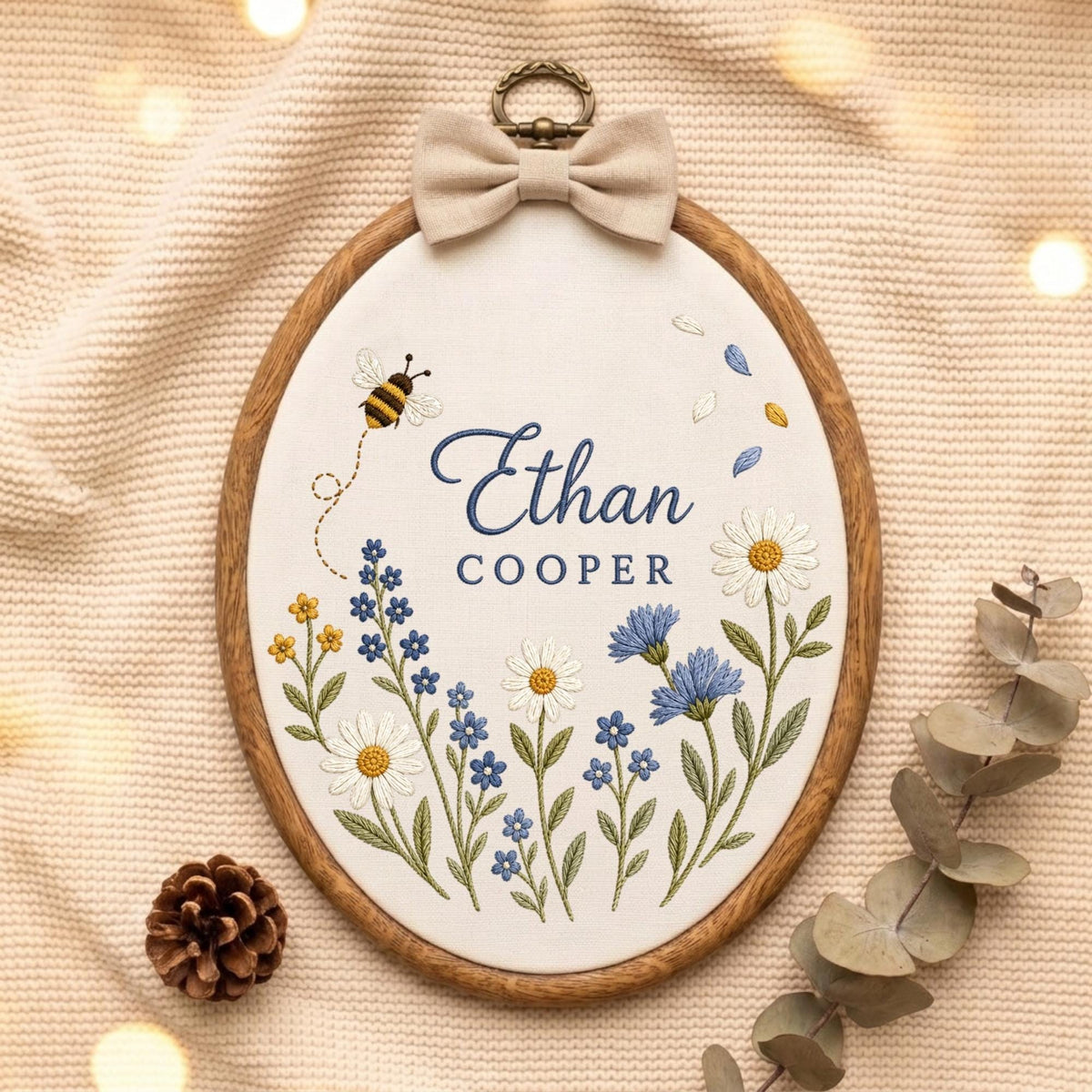 May include: An oval wooden embroidery hoop containing white fabric with the embroidered name Ethan Cooper in blue text. The design features a small bee, various blue and white wildflowers, and green stems. A beige fabric bow is attached to the top.