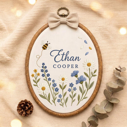 May include: An oval wooden embroidery hoop containing white fabric with the embroidered name Ethan Cooper in blue text. The design features a small bee, various blue and white wildflowers, and green stems. A beige fabric bow is attached to the top.