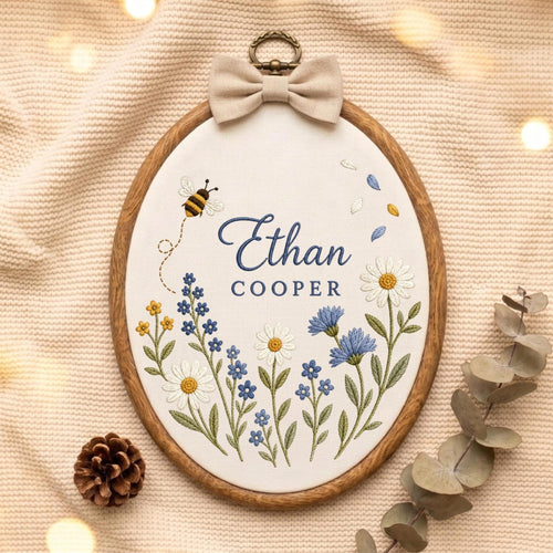May include: An oval wooden embroidery hoop containing white fabric with the embroidered name Ethan Cooper in blue text. The design features a small bee, various blue and white wildflowers, and green stems. A beige fabric bow is attached to the top.