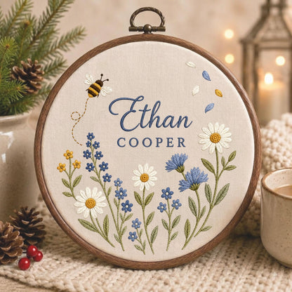 May include: A circular wooden embroidery hoop containing cream-colored fabric with hand-stitched floral designs, a small bee, and the text Ethan COOPER in dark blue thread. The embroidery features white daisies, blue cornflowers, and yellow buds.
