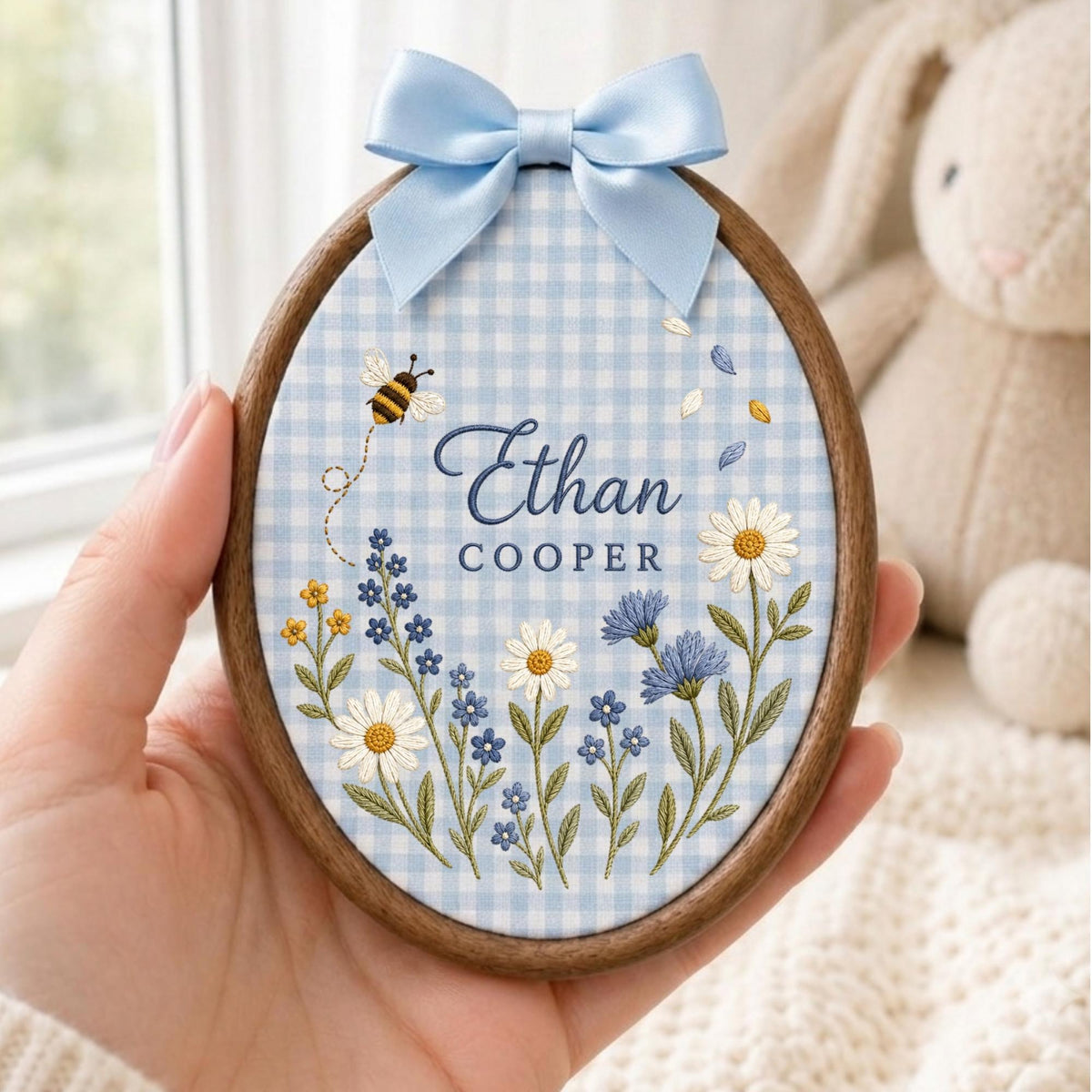 May include: An oval wooden embroidery hoop containing light blue and white gingham fabric. The fabric features embroidered wildflowers, a small bee, and the text Ethan COOPER in blue. A light blue satin ribbon bow is attached to the top.