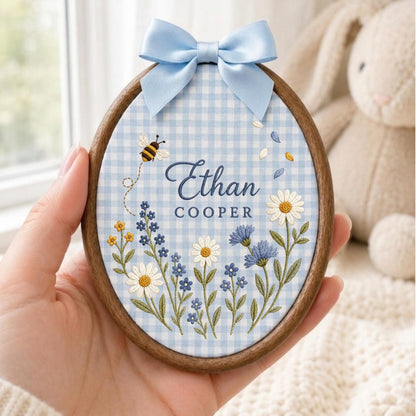 May include: An oval wooden embroidery hoop containing light blue and white gingham fabric. The fabric features embroidered wildflowers, a small bee, and the text Ethan COOPER in blue. A light blue satin ribbon bow is attached to the top.