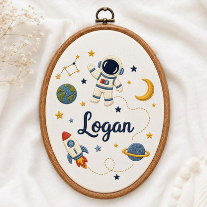 May include: An oval embroidery hoop containing white fabric with a space-themed design. The embroidery features an astronaut, a rocket, the Earth, a planet with rings, a crescent moon, and stars. The name Logan is stitched in dark blue script.