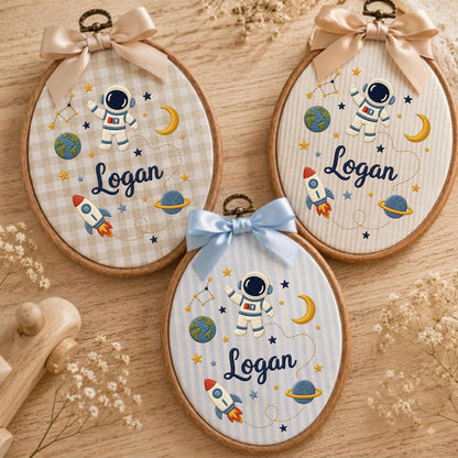 May include: Three oval wooden embroidery hoops featuring fabric backgrounds with space-themed embroidery including an astronaut, rocket, planet, moon, and stars. Each hoop displays the name Logan in dark blue script and has a decorative satin bow.