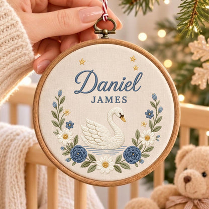 May include: A circular wooden embroidery hoop containing a cream-colored fabric panel. The fabric features an embroidered white swan centered above blue and white floral wreaths, with the blue text Daniel JAMES and small gold stars above.