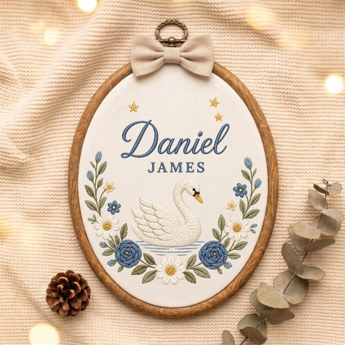 May include: An oval wooden embroidery hoop containing white fabric with the embroidered name Daniel JAMES in blue, a white swan, blue and white flowers, and small gold stars. A beige fabric bow is attached to the top metal hanging ring.