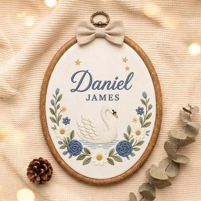 May include: An oval wooden embroidery hoop containing white fabric with the embroidered name Daniel JAMES in blue, a white swan, blue and white flowers, and small gold stars. A beige fabric bow is attached to the top metal hanging ring.