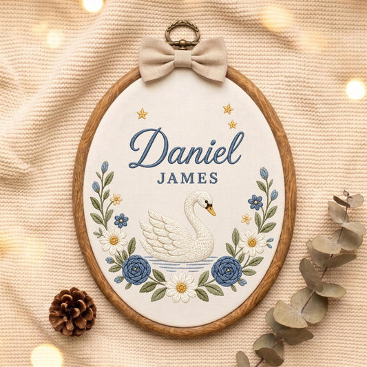 May include: An oval wooden embroidery hoop containing white fabric with the embroidered name Daniel JAMES in blue, a white swan, blue and white flowers, and small gold stars. A beige fabric bow is attached to the top metal hanging ring.