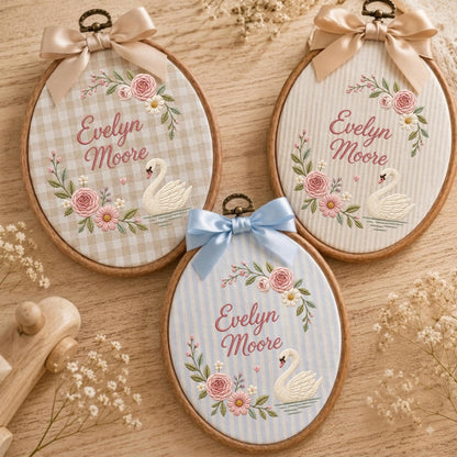 May include: Three oval wooden embroidery hoops featuring fabric backgrounds with gingham or striped patterns. Each hoop displays embroidered pink flowers, green leaves, a white swan, and the name Evelyn Moore in script, topped with a satin bow.
