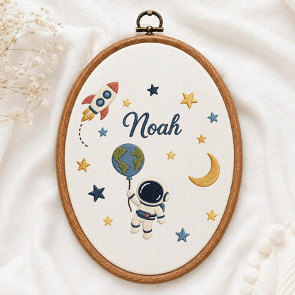 May include: An oval embroidery hoop containing white fabric with a space-themed design. The embroidery features a rocket, a crescent moon, stars, an astronaut holding a globe balloon, and the name Noah in dark blue cursive script.