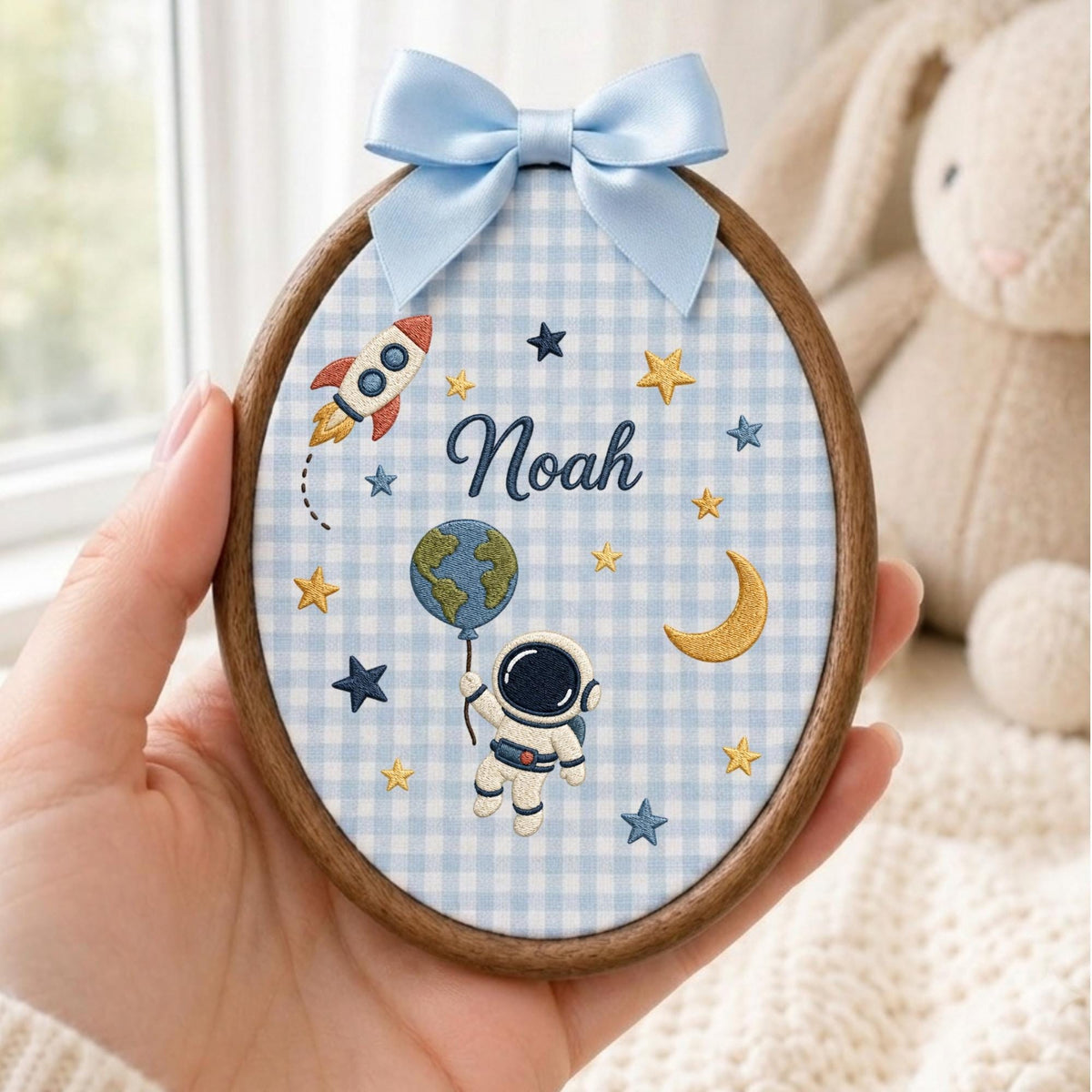 May include: An oval wooden embroidery hoop containing light blue and white gingham fabric. The fabric features embroidered space motifs including a rocket, stars, a crescent moon, an astronaut holding a globe, and the name Noah in dark blue script.