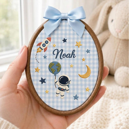 May include: An oval wooden embroidery hoop containing light blue and white gingham fabric. The fabric features embroidered space motifs including a rocket, stars, a crescent moon, an astronaut holding a globe, and the name Noah in dark blue script.