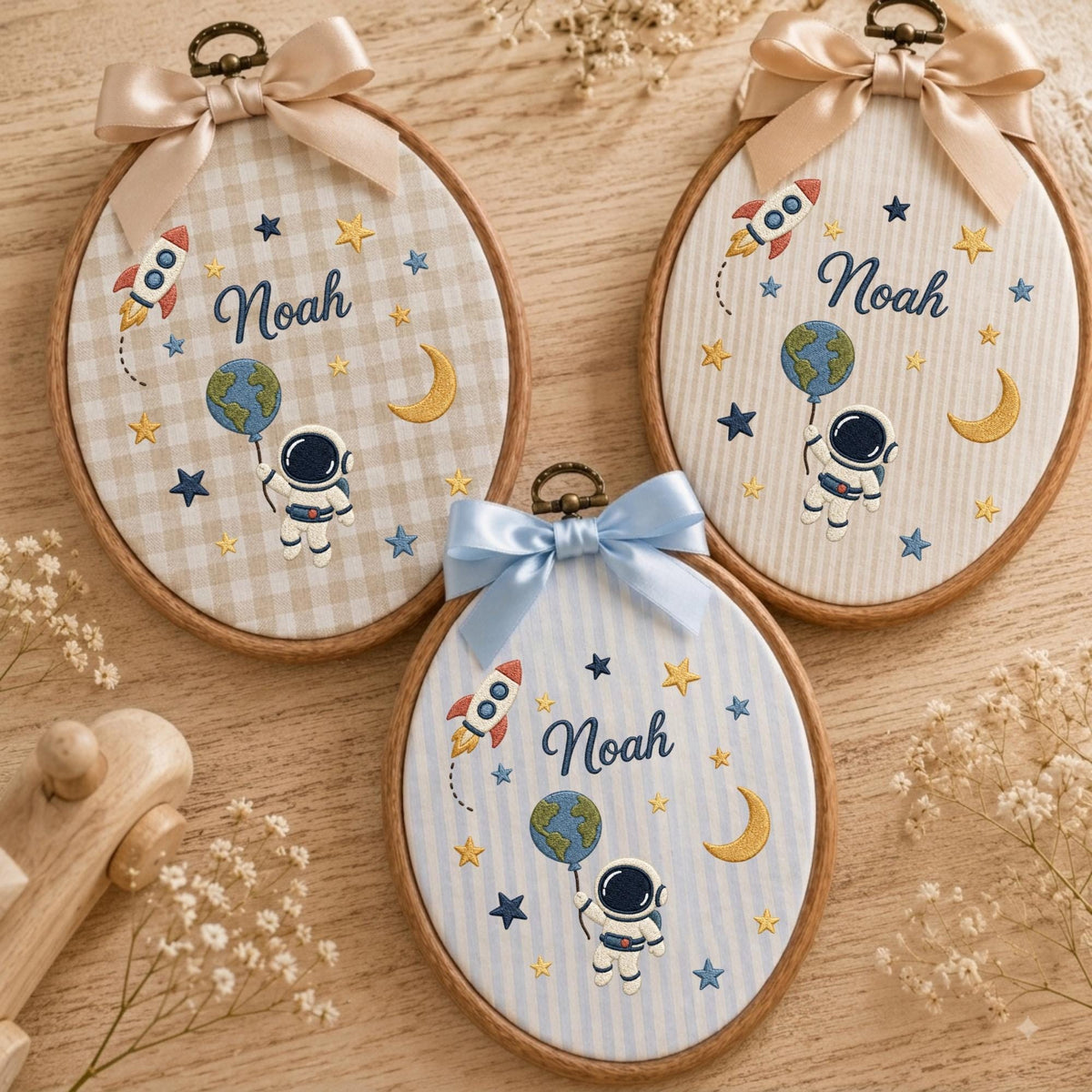May include: Three oval embroidery hoops featuring space-themed designs with the name Noah stitched in blue. Each hoop displays an astronaut holding a globe balloon, a rocket, stars, and a crescent moon, topped with a decorative satin ribbon bow.