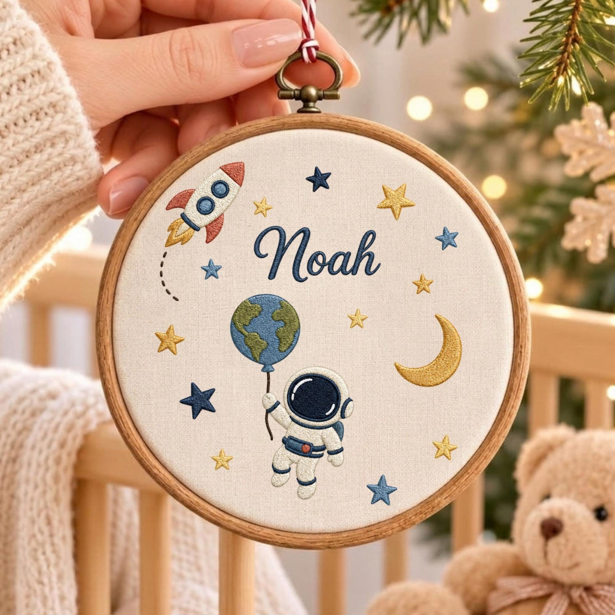 May include: A round wooden embroidery hoop containing a cream-colored fabric panel with space-themed embroidery. The design features the name Noah in blue script, a rocket ship, a crescent moon, stars, and an astronaut holding a globe balloon.