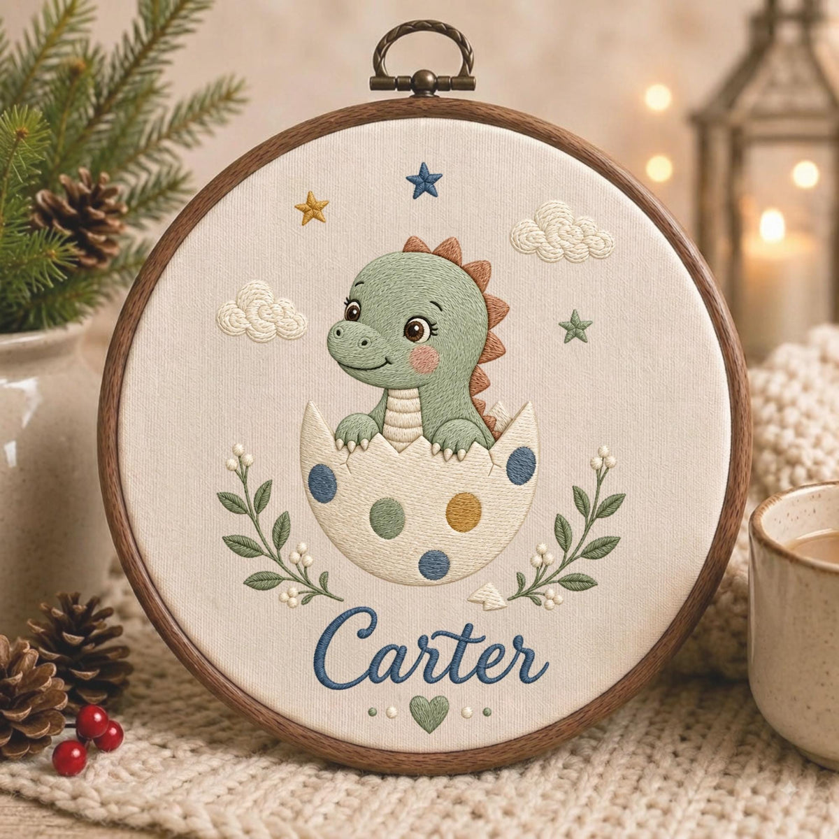 May include: A circular wooden embroidery hoop containing a cream-colored fabric with a stitched design of a green dinosaur hatching from a spotted egg. The embroidery includes clouds, stars, green leafy branches, and the name Carter in blue script.