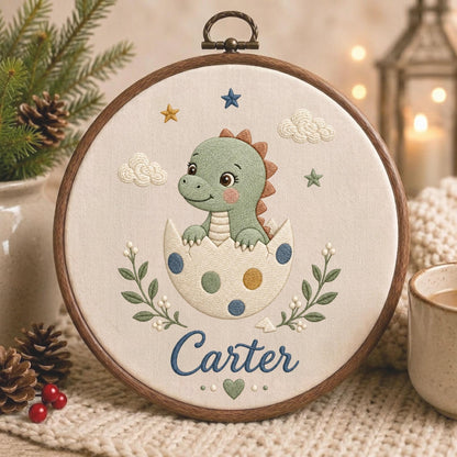 May include: A circular wooden embroidery hoop containing a cream-colored fabric with a stitched design of a green dinosaur hatching from a spotted egg. The embroidery includes clouds, stars, green leafy branches, and the name Carter in blue script.