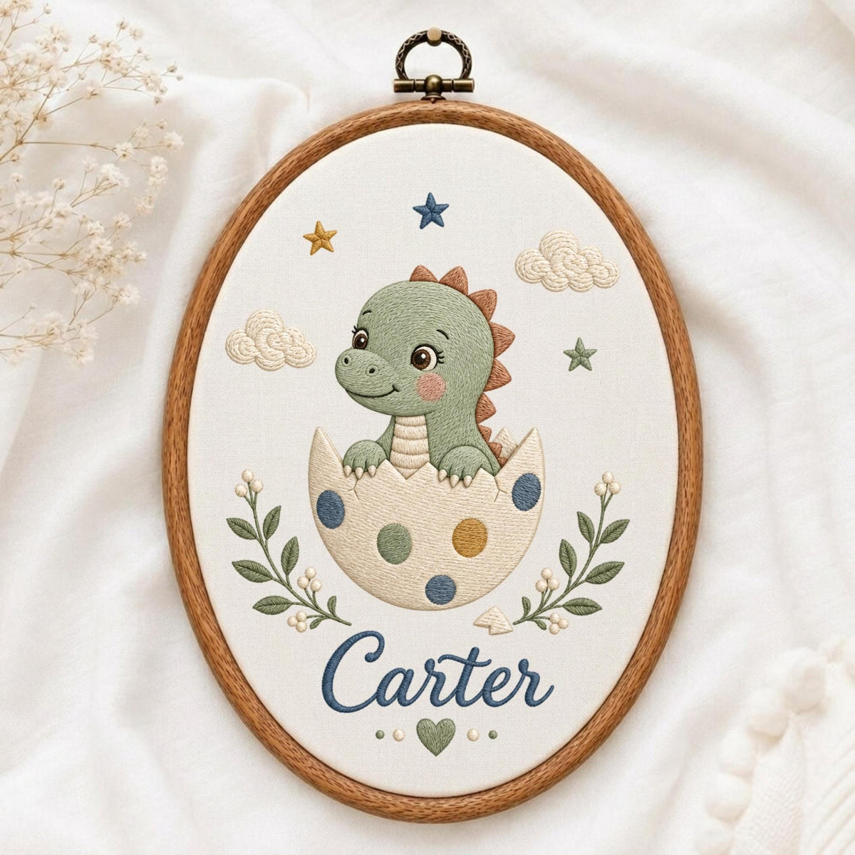 May include: An oval embroidery hoop containing a fabric panel with a stitched green dinosaur hatching from a polka-dot egg. The design includes embroidered clouds, stars, and leafy branches surrounding the name Carter in blue script lettering.