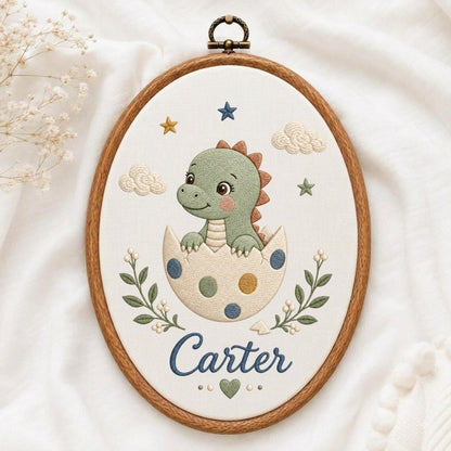 May include: An oval embroidery hoop containing a fabric panel with a stitched green dinosaur hatching from a polka-dot egg. The design includes embroidered clouds, stars, and leafy branches surrounding the name Carter in blue script lettering.