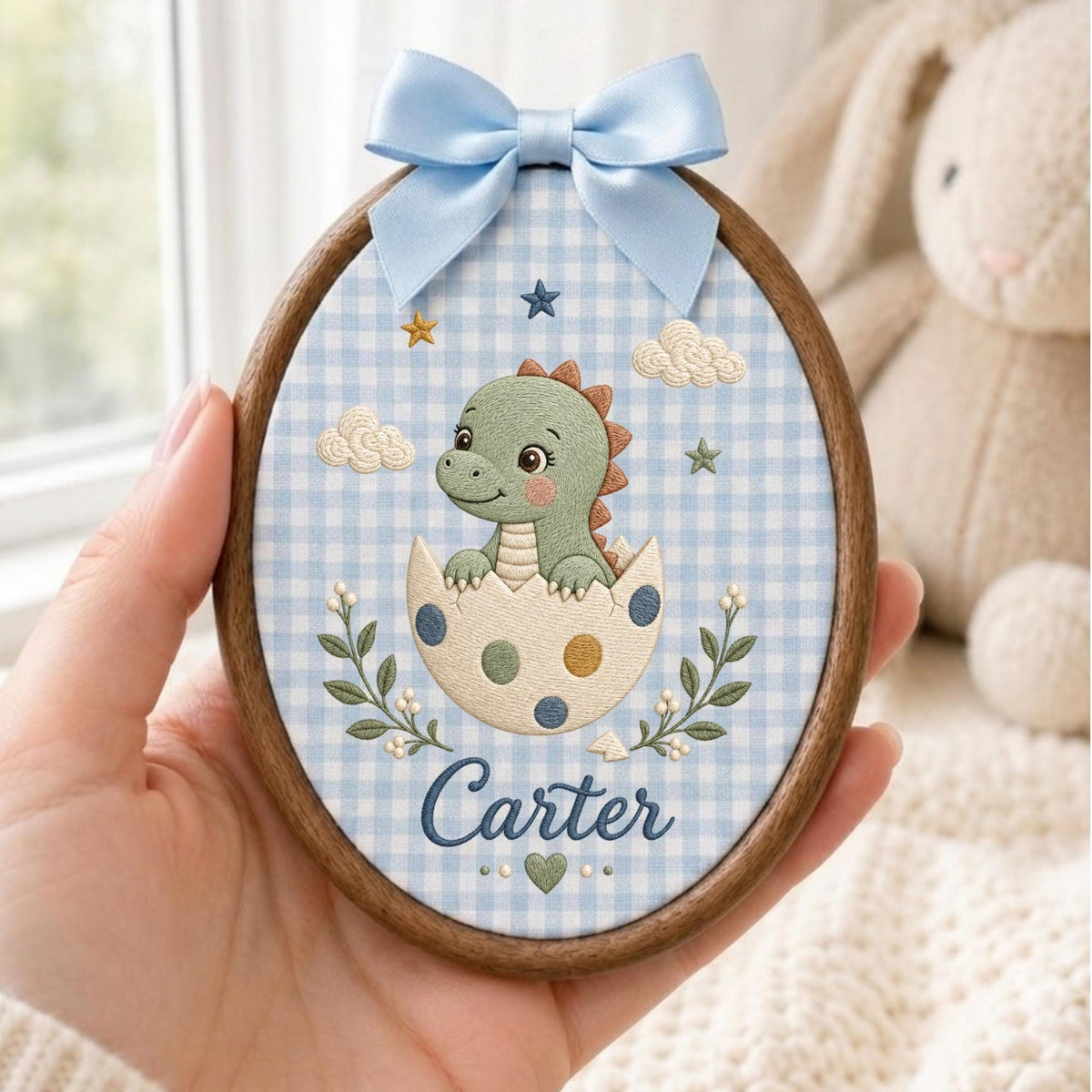 May include: An oval embroidery hoop featuring a light blue and white gingham fabric background. The design includes a green baby dinosaur hatching from a spotted egg, surrounded by embroidered clouds, stars, and leaves. The name Carter is stitched in blue script at the bottom, topped with a light blue satin bow.