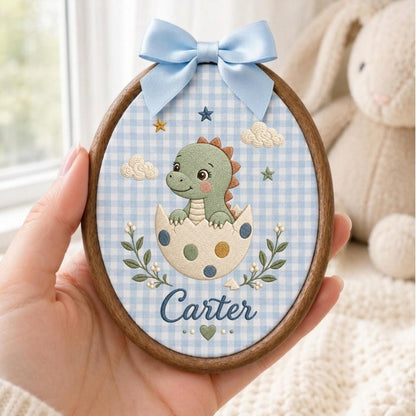 May include: An oval embroidery hoop featuring a light blue and white gingham fabric background. The design includes a green baby dinosaur hatching from a spotted egg, surrounded by embroidered clouds, stars, and leaves. The name Carter is stitched in blue script at the bottom, topped with a light blue satin bow.