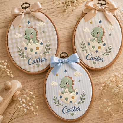 May include: Three oval wooden embroidery hoops featuring fabric backgrounds with embroidered green dinosaurs hatching from spotted eggs. Each hoop is topped with a satin ribbon bow and displays the name Carter in blue script embroidery.