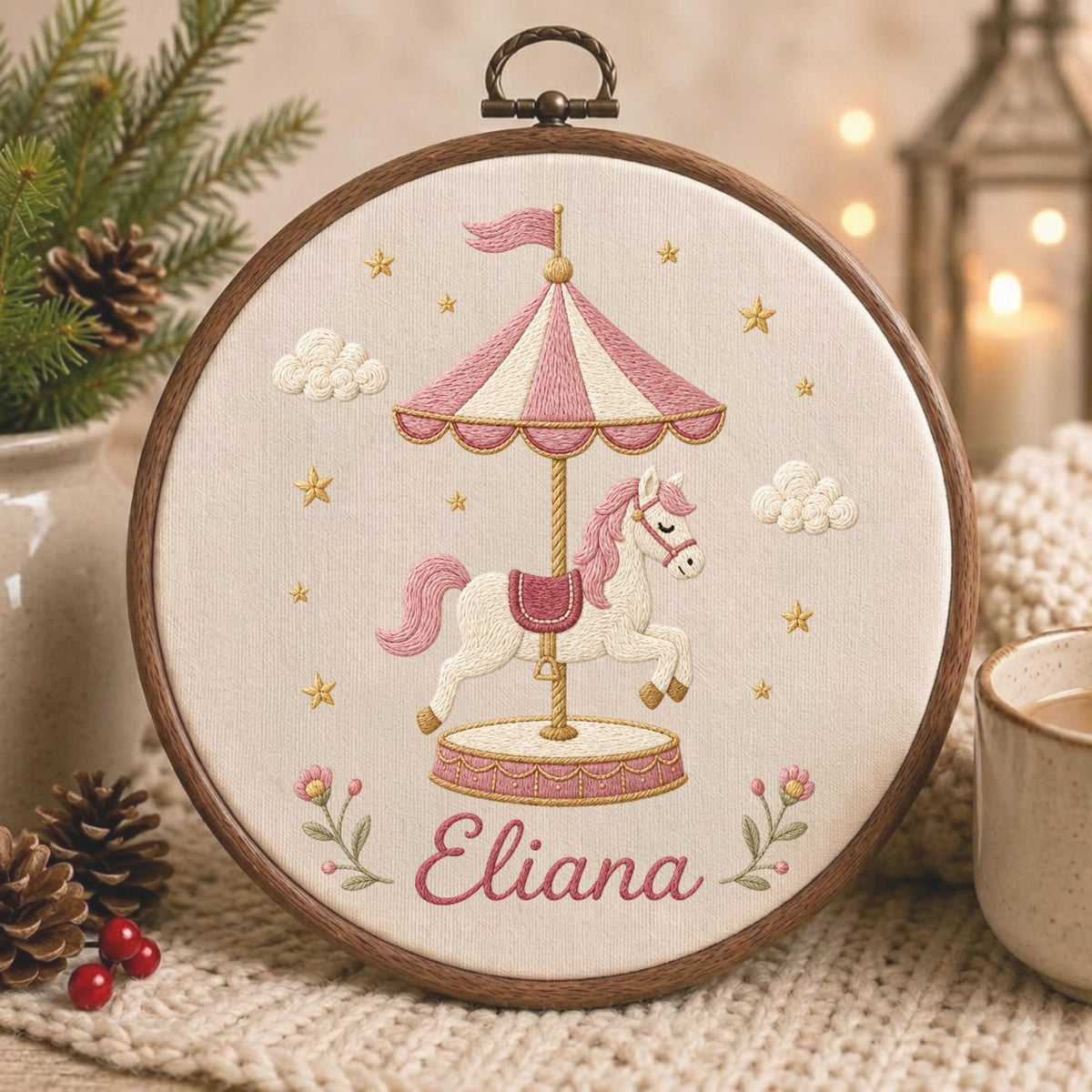 May include: A circular wooden embroidery hoop containing a cream-colored fabric panel. The embroidery features a carousel with a white horse, a pink and white striped canopy, and small gold stars. The name Eliana is stitched in pink cursive.