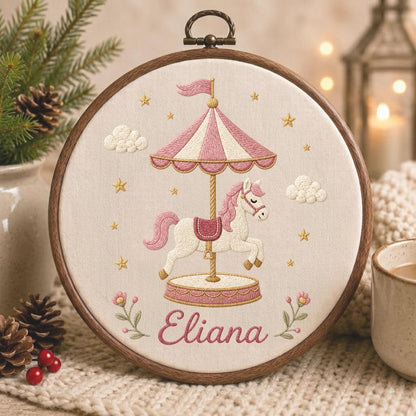 May include: A circular wooden embroidery hoop containing a cream-colored fabric panel. The embroidery features a carousel with a white horse, a pink and white striped canopy, and small gold stars. The name Eliana is stitched in pink cursive.