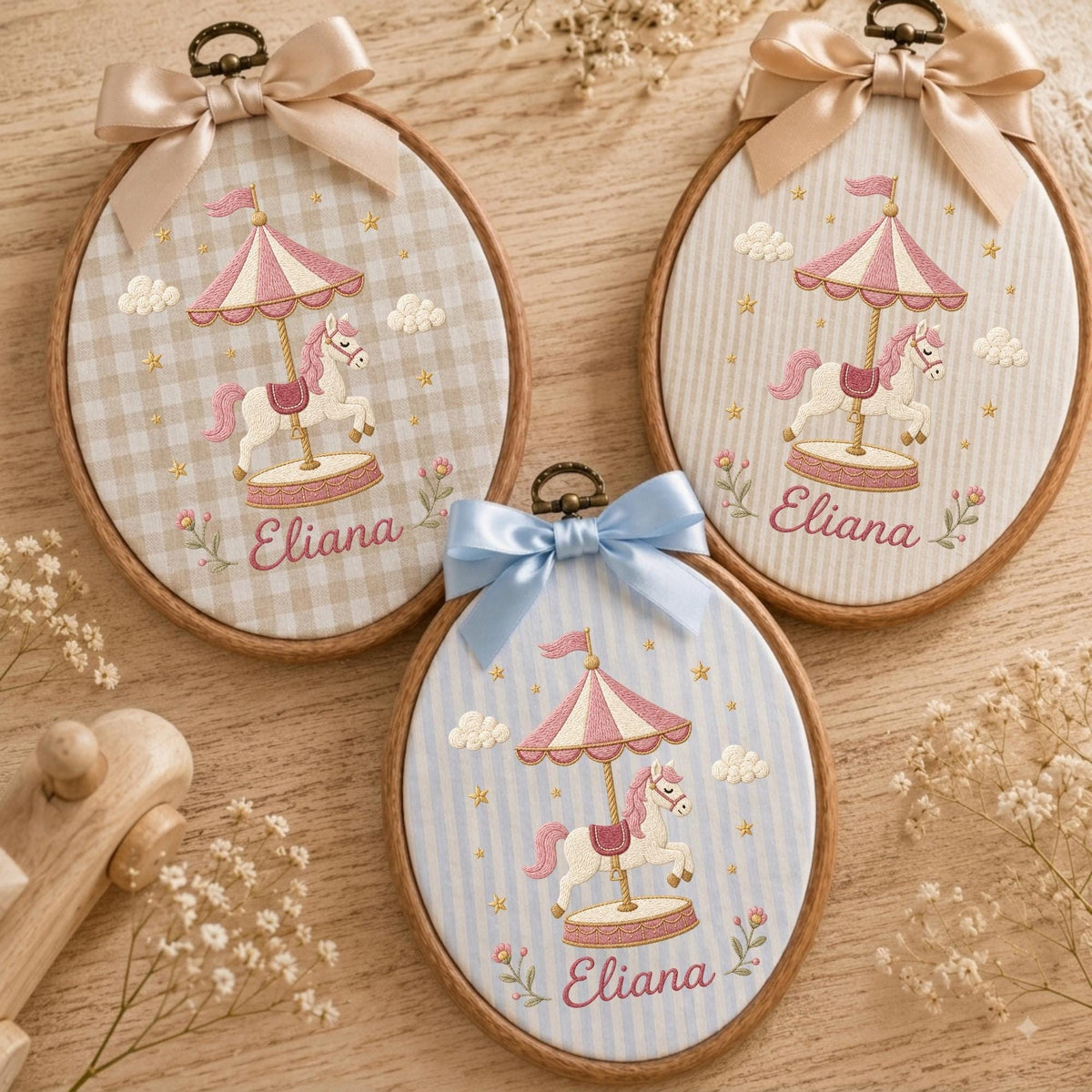 May include: Three oval wooden embroidery hoops featuring fabric with carousel horse designs. Each hoop displays a pink and white carousel, a white horse, and the name Eliana embroidered in pink. The hoops are topped with satin bows in beige or blue.