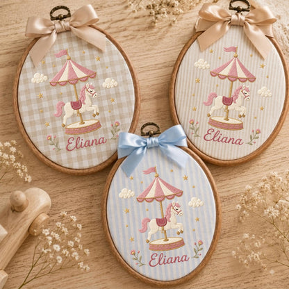 May include: Three oval wooden embroidery hoops featuring fabric with carousel horse designs. Each hoop displays a pink and white carousel, a white horse, and the name Eliana embroidered in pink. The hoops are topped with satin bows in beige or blue.