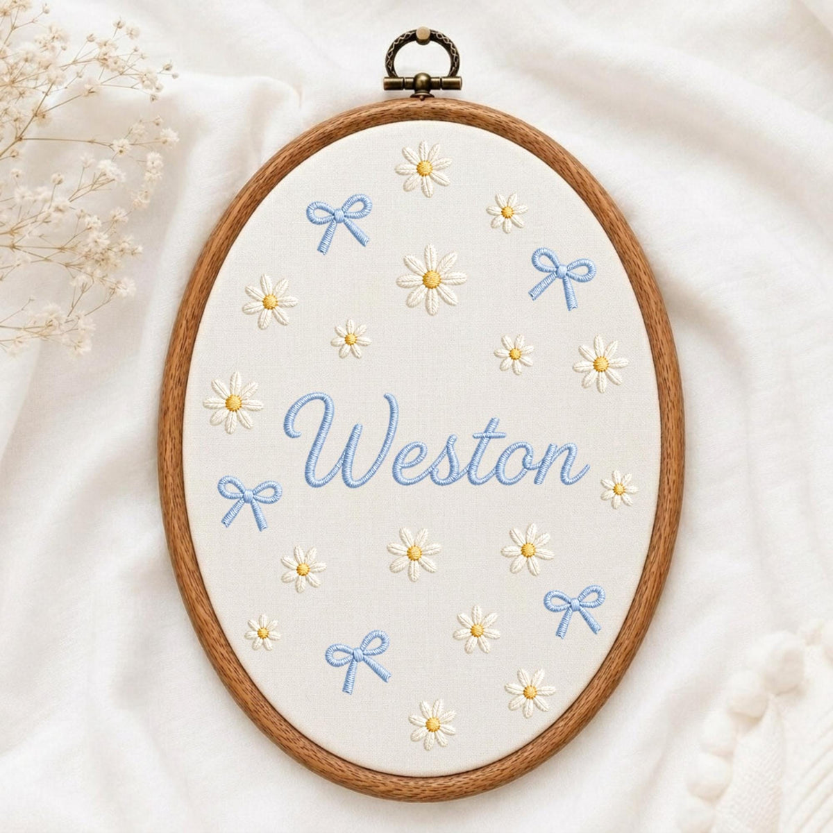 May include: An oval wooden embroidery hoop containing white fabric with the name Weston stitched in light blue script. The fabric is decorated with embroidered white daisies with yellow centers and small light blue bows scattered throughout.