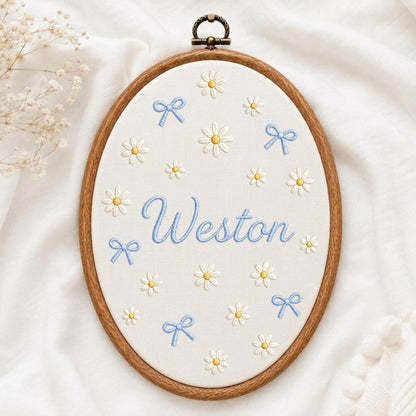 May include: An oval wooden embroidery hoop containing white fabric with the name Weston stitched in light blue script. The fabric is decorated with embroidered white daisies with yellow centers and small light blue bows scattered throughout.