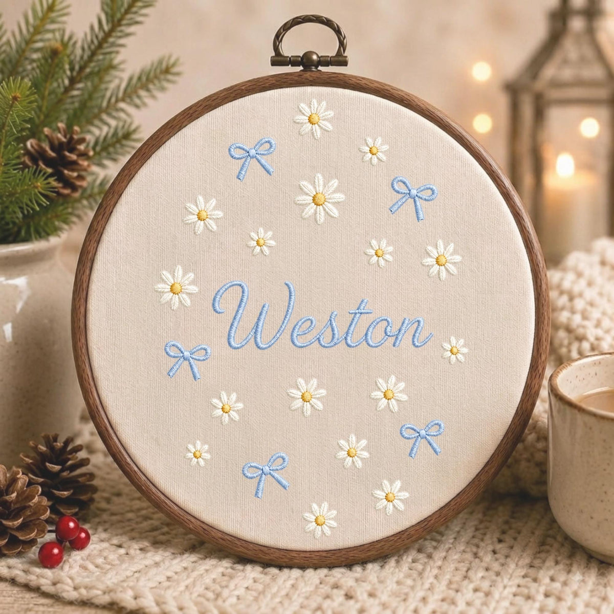 May include: A circular wooden embroidery hoop containing beige fabric with embroidered white daisies, light blue bows, and the name Weston stitched in light blue cursive script in the center. The hoop features a metal hanging loop at the top.