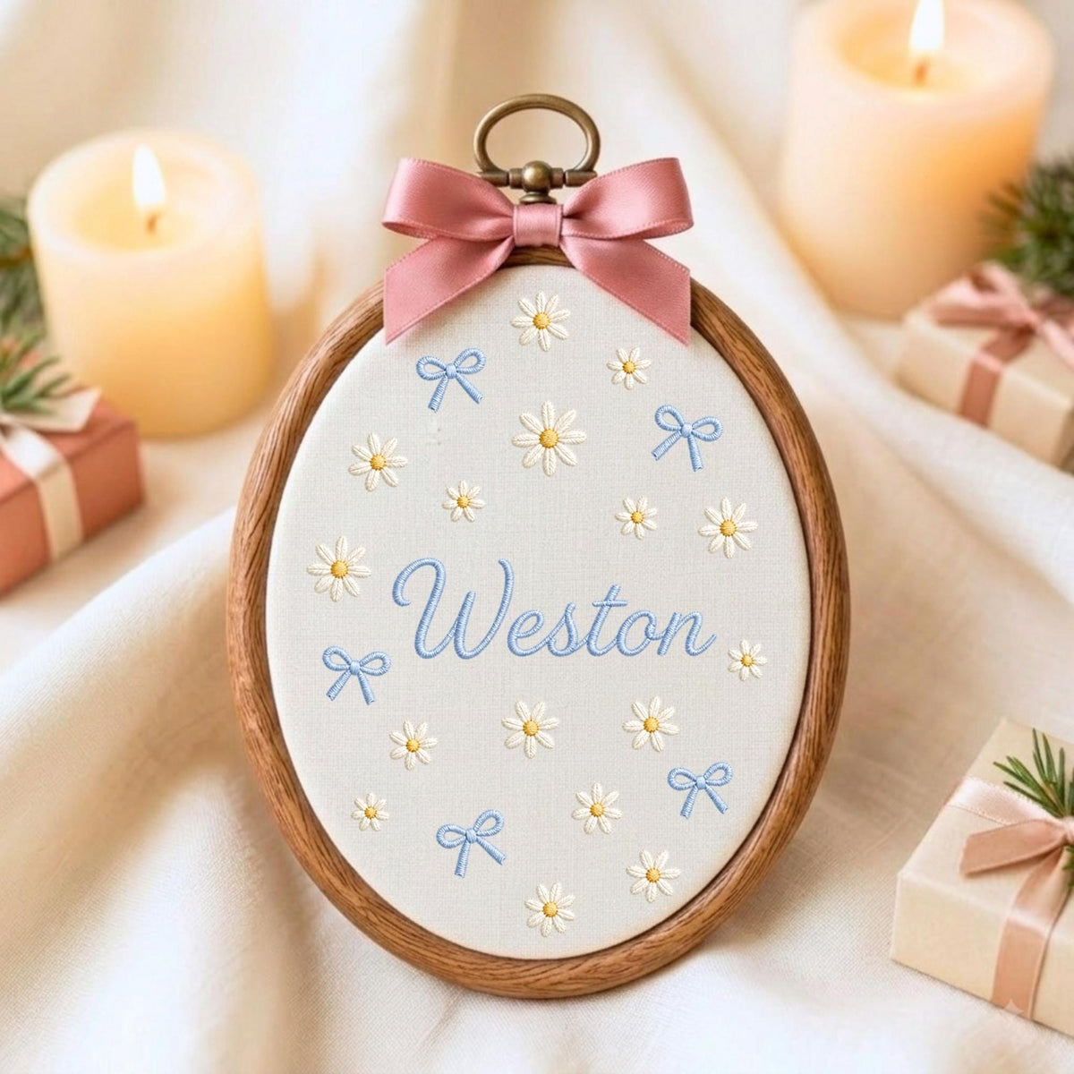 May include: An oval wooden embroidery hoop containing off-white fabric with embroidered white daisies, light blue bows, and the name Weston in blue script. A dusty pink satin ribbon bow is attached to the top metal hanging loop.
