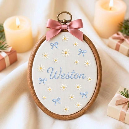 May include: An oval wooden embroidery hoop containing off-white fabric with embroidered white daisies, light blue bows, and the name Weston in blue script. A dusty pink satin ribbon bow is attached to the top metal hanging loop.