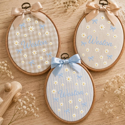 May include: Three oval wooden embroidery hoops featuring fabric backgrounds with embroidered white daisies, light blue bows, and the name Weston in blue script. Each hoop is topped with a decorative satin ribbon bow in beige or light blue.