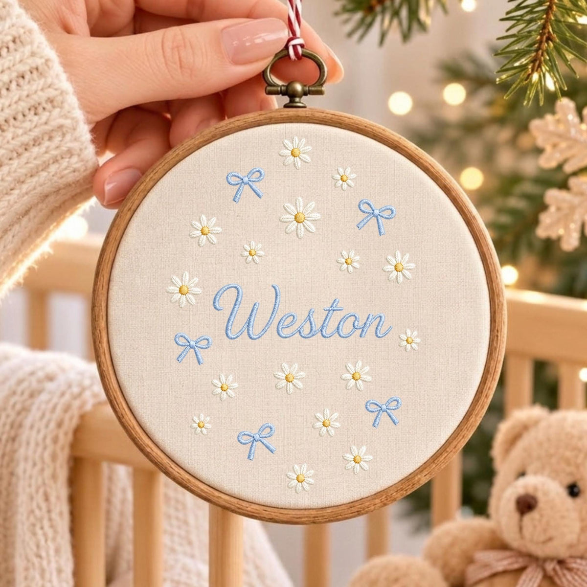 May include: A circular wooden embroidery hoop containing beige fabric with the name Weston embroidered in blue script. The fabric features a pattern of small white daisies with yellow centers and light blue bows scattered around the central name.