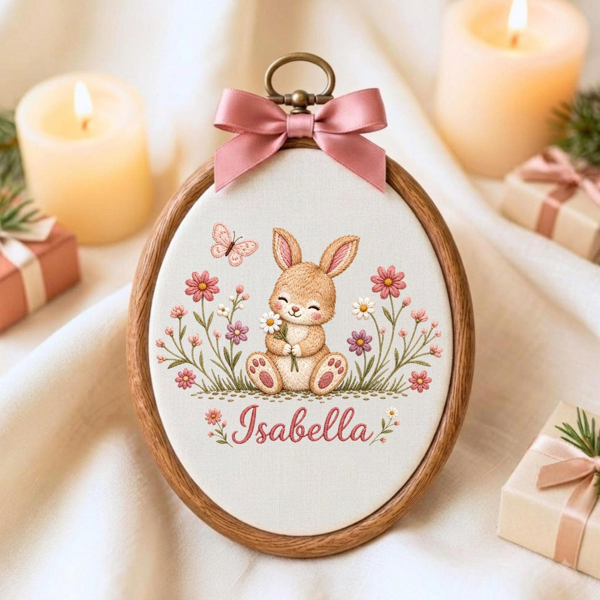 May include: An oval wooden embroidery hoop containing a fabric panel with a stitched illustration of a brown bunny holding a white daisy, surrounded by pink and purple flowers and a butterfly. The name Isabella is embroidered in pink script below.
