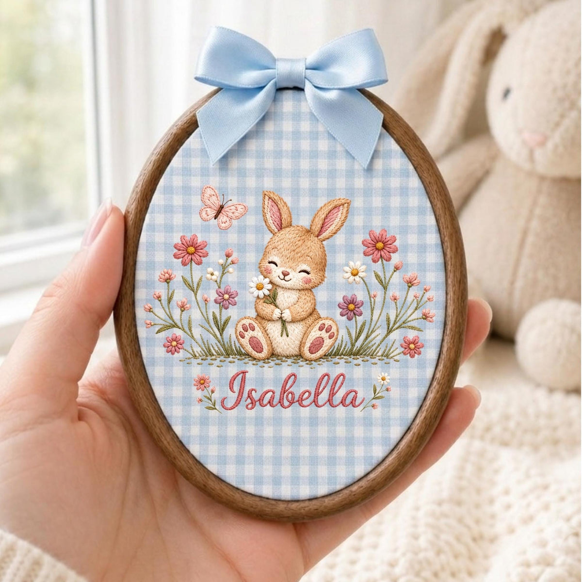 May include: An oval wooden embroidery hoop frame containing fabric with a light blue gingham pattern. The fabric features a printed illustration of a brown bunny holding a white flower, surrounded by pink flowers and a butterfly, with the name Isabella.
