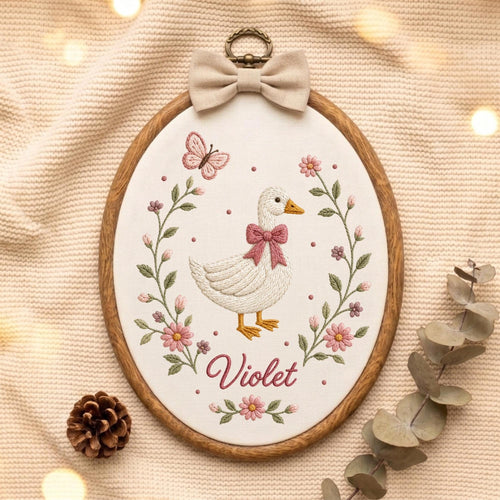 May include: An oval embroidery hoop containing a white fabric panel with a stitched white duck wearing a pink bow. The duck is surrounded by a floral wreath with pink flowers, green leaves, and a butterfly. The name Violet is stitched in pink.