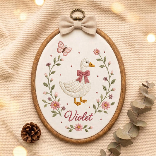 May include: An oval embroidery hoop containing a white fabric panel with a stitched white duck wearing a pink bow. The duck is surrounded by a floral wreath with pink flowers, green leaves, and a butterfly. The name Violet is stitched in pink.