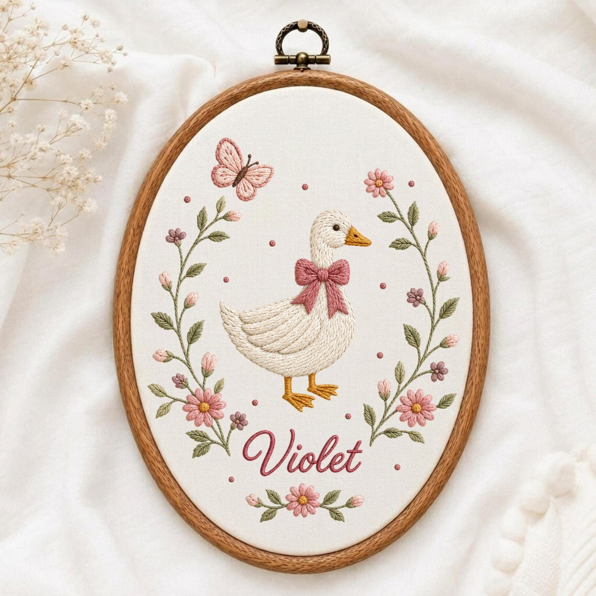 May include: An oval wooden embroidery hoop containing white fabric with a hand-stitched design of a white goose wearing a pink bow, surrounded by a floral wreath of pink flowers and green leaves, a pink butterfly, and the name Violet in pink script.