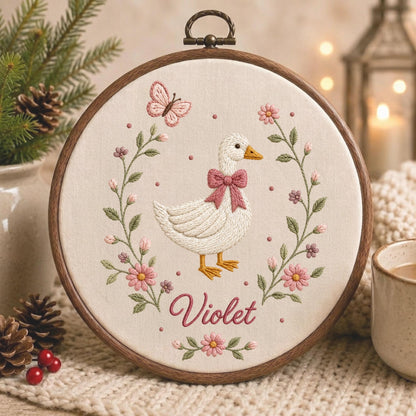 May include: A circular wooden embroidery hoop containing a cream-colored fabric panel. The embroidery features a white duck with a pink bow, surrounded by a floral wreath of pink flowers, green leaves, and a butterfly, with the name Violet below.