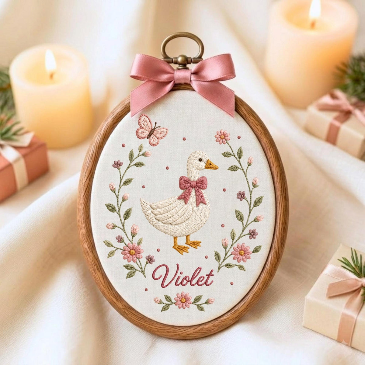 May include: An oval wooden embroidery hoop containing white fabric with a stitched design of a white goose wearing a pink bow, surrounded by pink flowers and a butterfly. The name Violet is embroidered in pink below the goose, and a pink satin bow is attached to the top.