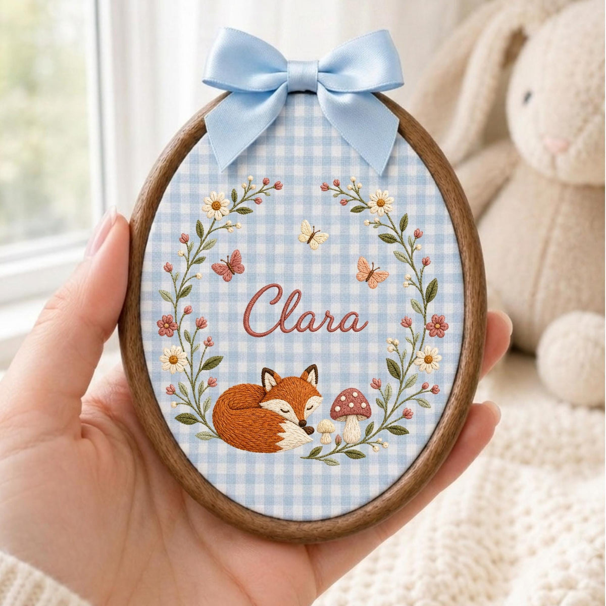 May include: An oval wooden embroidery hoop featuring a light blue and white gingham fabric background. The embroidery depicts a sleeping fox, a mushroom, and a floral wreath with butterflies. The name Clara is stitched in the center with a blue bow.