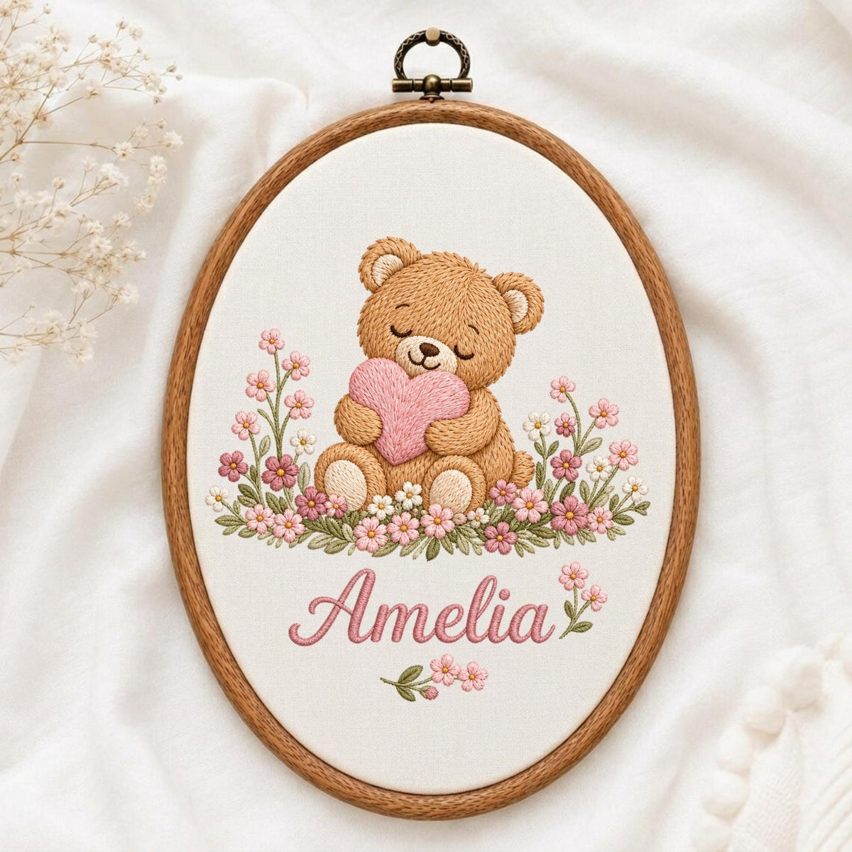 May include: An oval wooden embroidery hoop containing a fabric panel with a stitched design of a brown teddy bear holding a pink heart, surrounded by pink and white flowers. The name Amelia is embroidered in pink script below the bear.